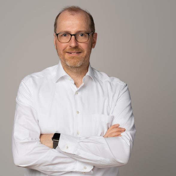 Christian Meiners Mann mit Brille trägt ein weißes Hemd, steht mit verschränkten Armen vor neutralem Hintergrund.
